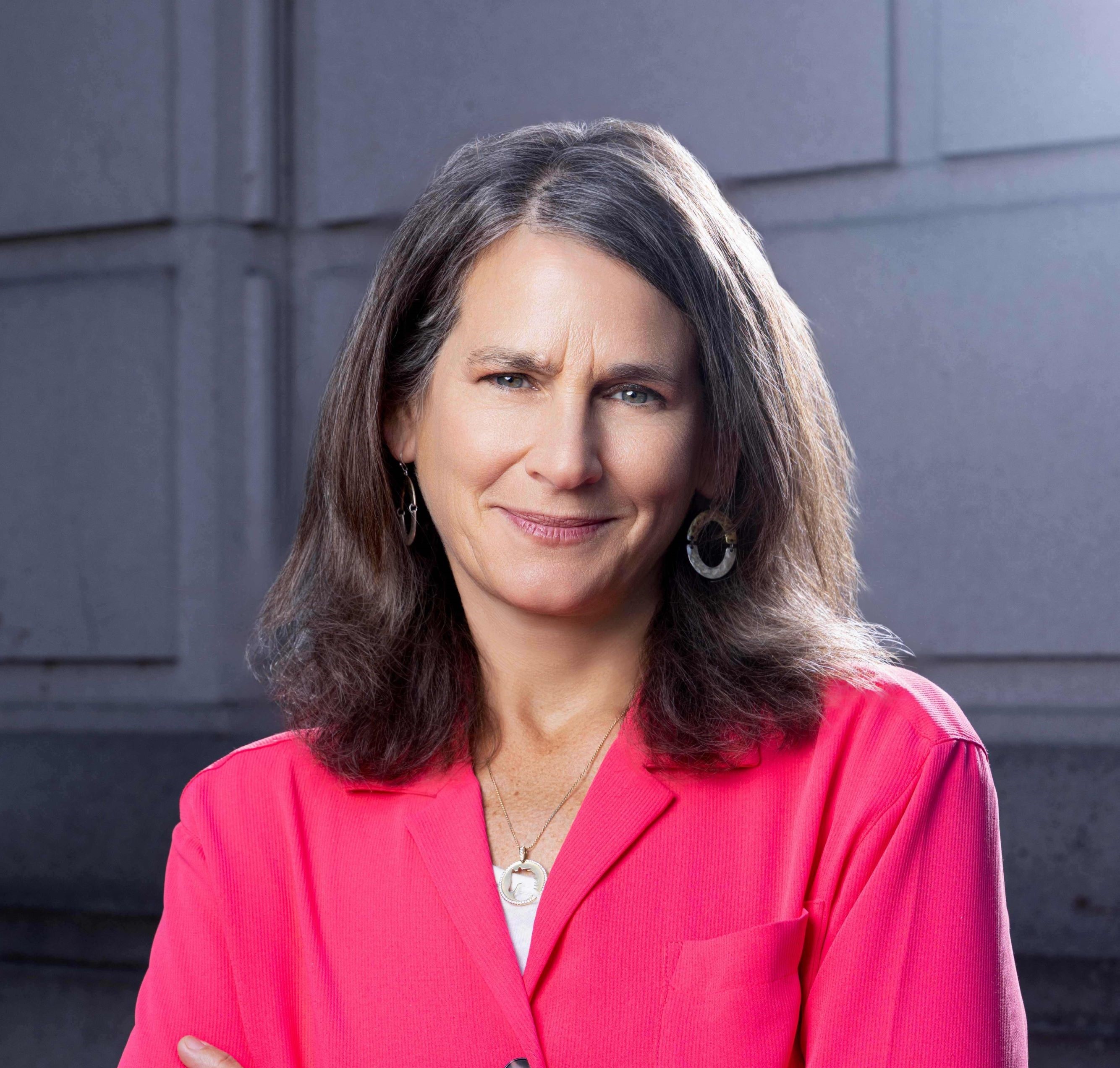 Woman with dark long hair wearing a bright pink jacket while standing in front of a building