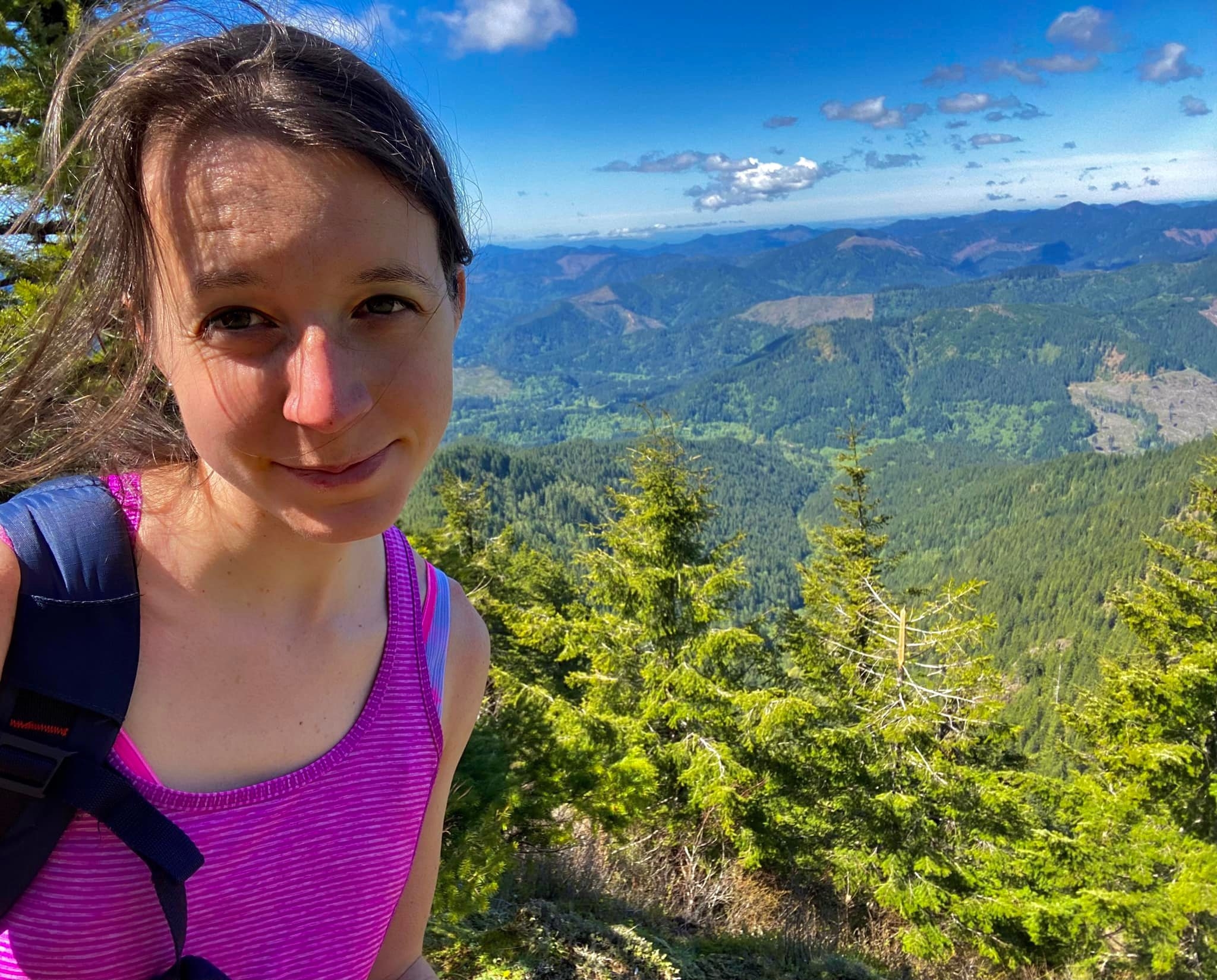Woman taking selfie atop on overlook on a sunny day. Trees and mostly blue skies can be seen behind her.