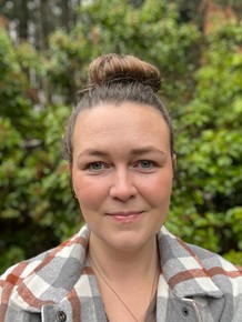 Woman wearing a plaid jacket and her hair in a bun, standing in front of a green bush.