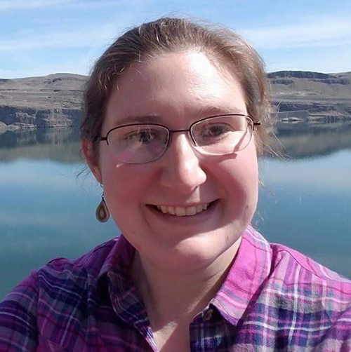 Woman wearing purple plaid shirt standing posing for a selfie. A body of water and mountains can be seen behind her.