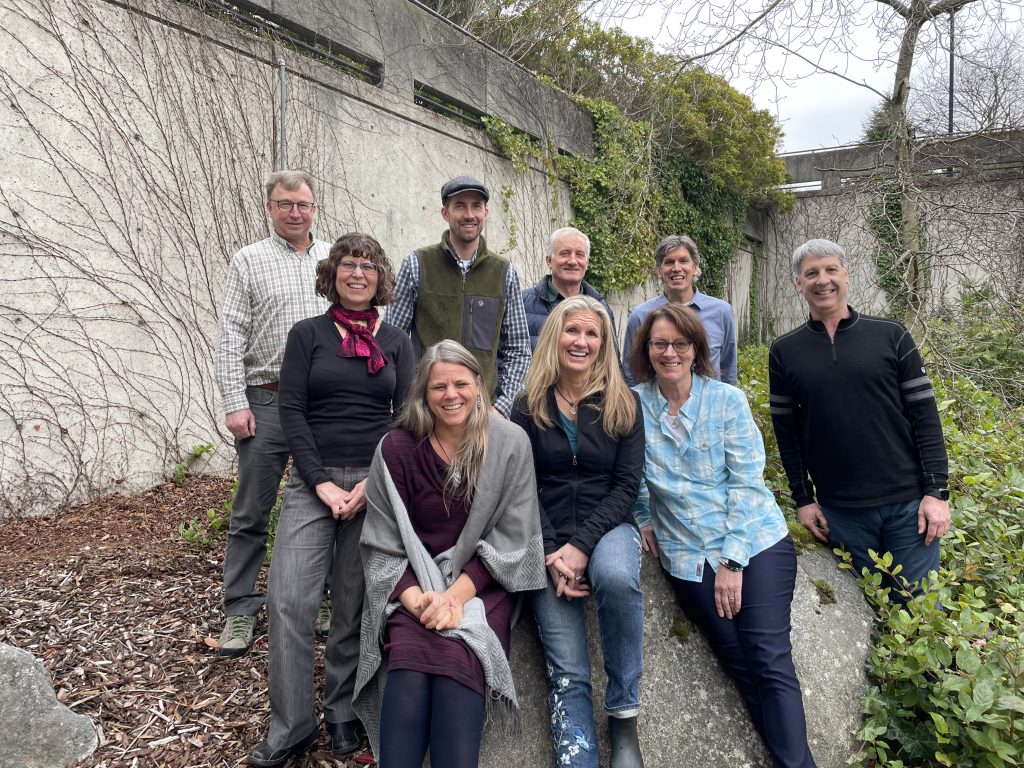 Nine of the 10 Review Panel members posing for a photo. A few are seated upon a large rock. Others are flanking behind them and to the sides. A large wall can be seen behind the group.