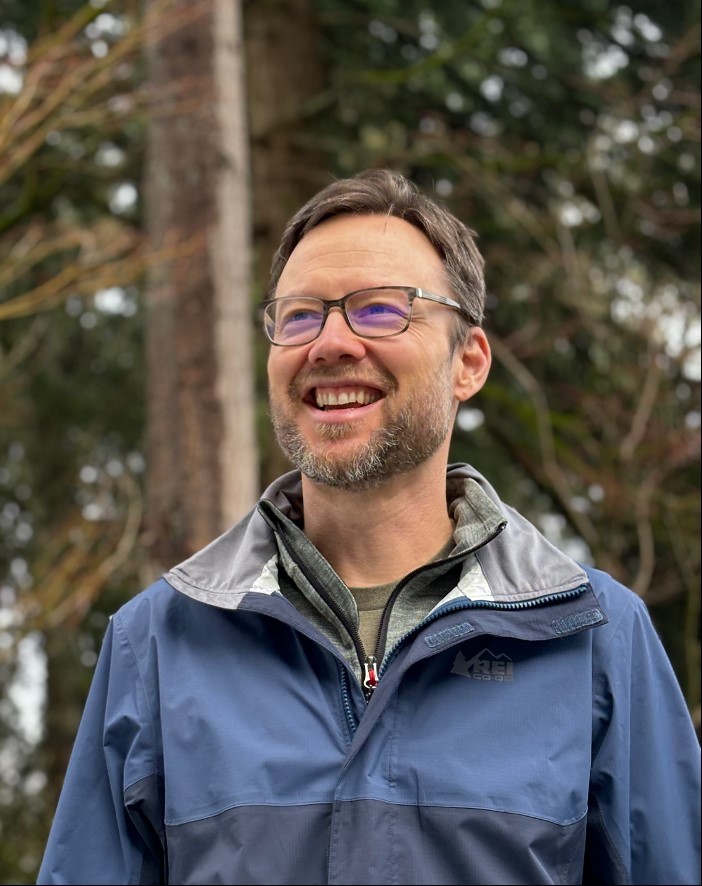 Man wearing glasses standing in front of trees.