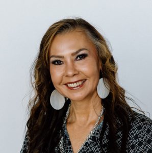 A woman with long dark hair and round white earrings smiling at camera