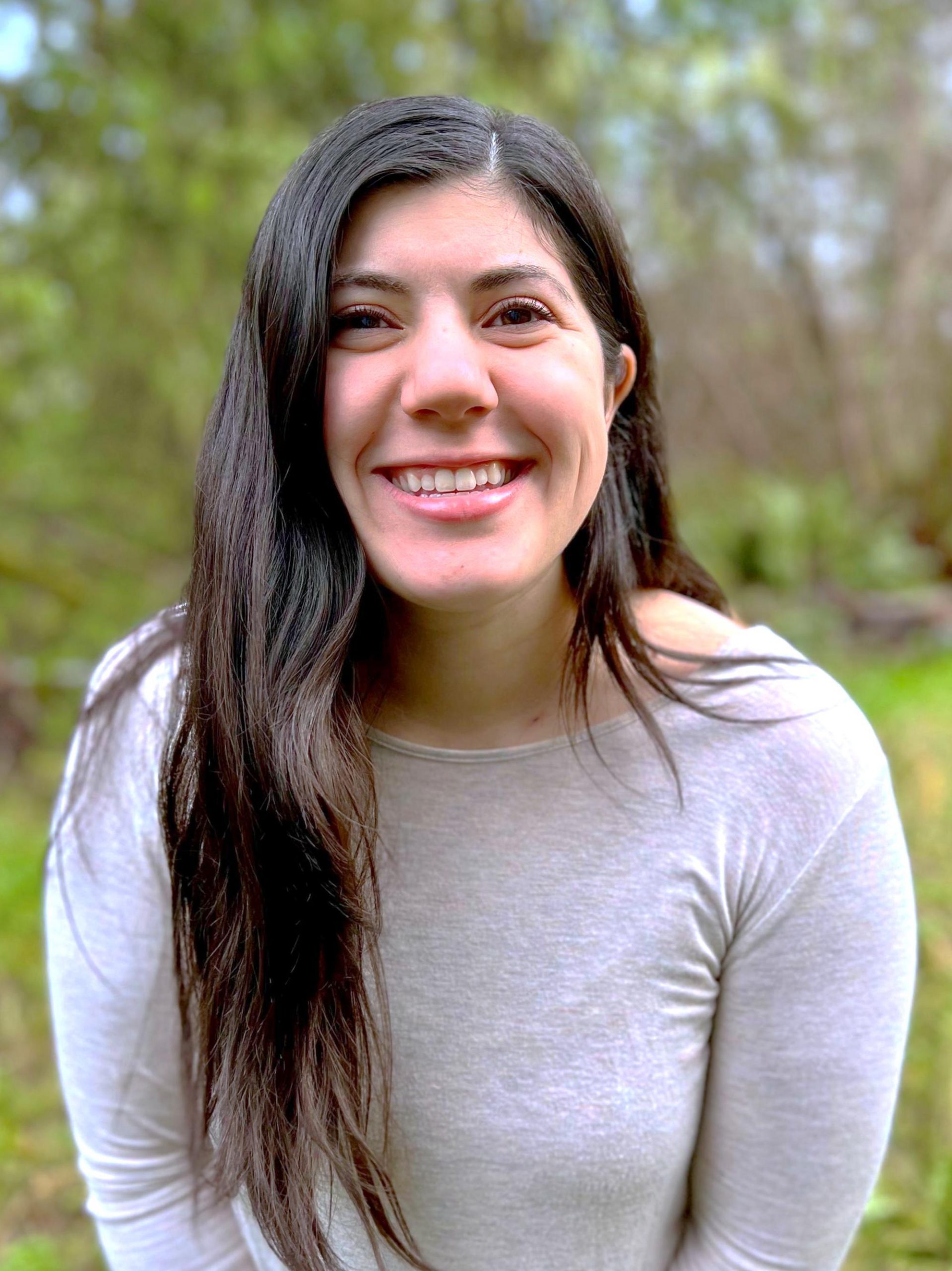 Dark haired woman in a white long-sleeved shirt smiling with green grass and trees in the background