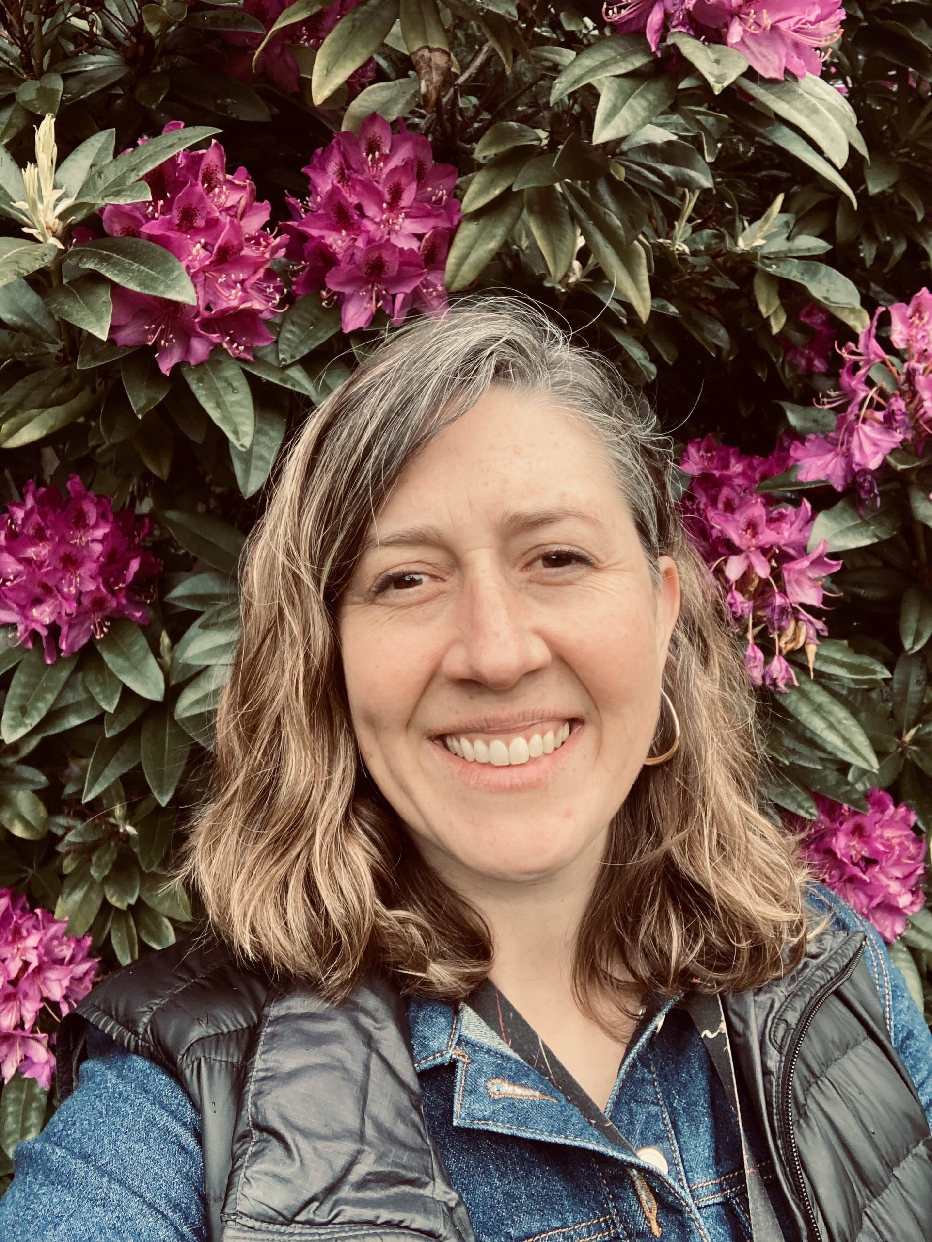 Woman smiling while standing in front of a tall bush with green leaves and pinkish purple flowers